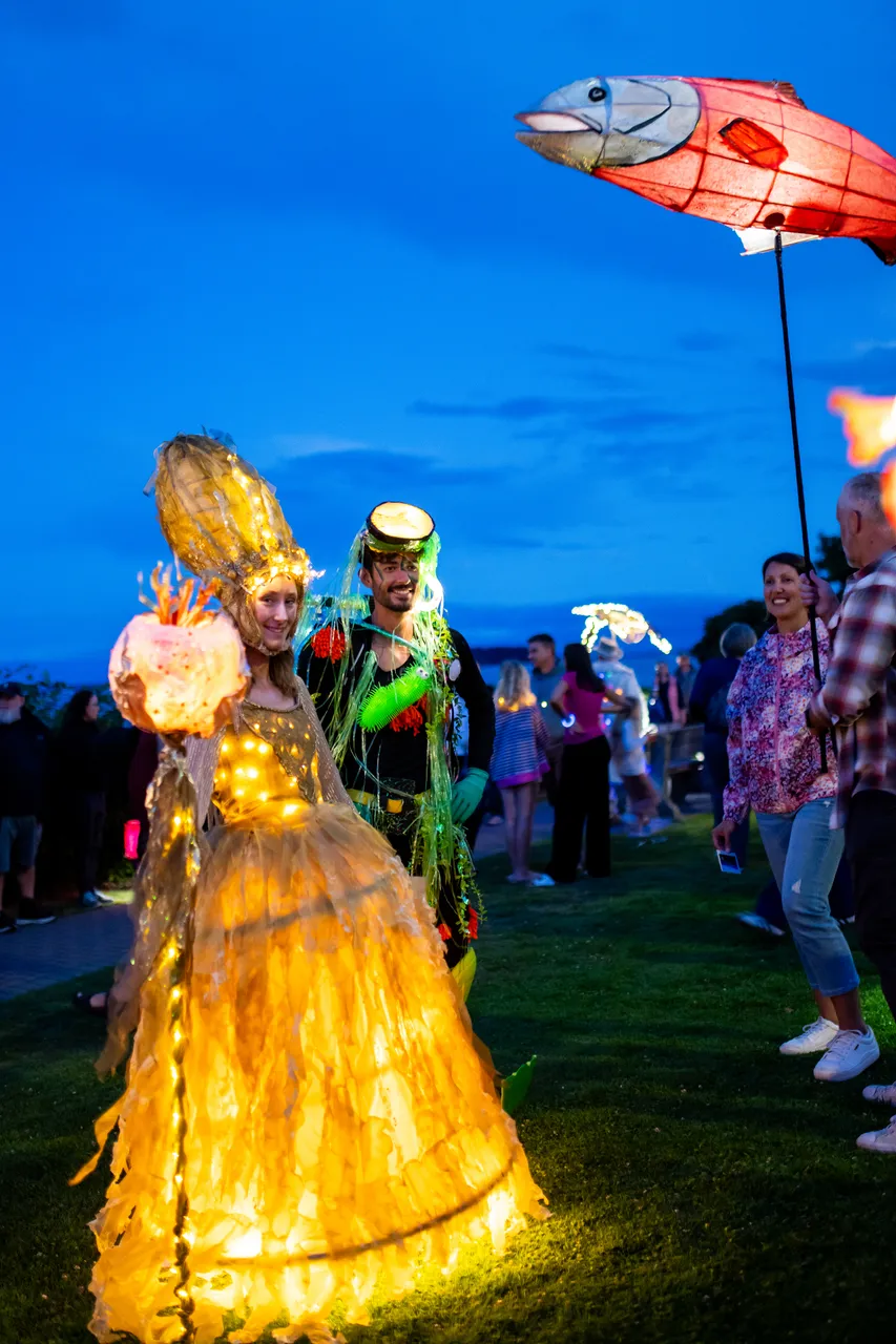 Performers in illuminated lantern gowns lead a crowd along a street at twilight